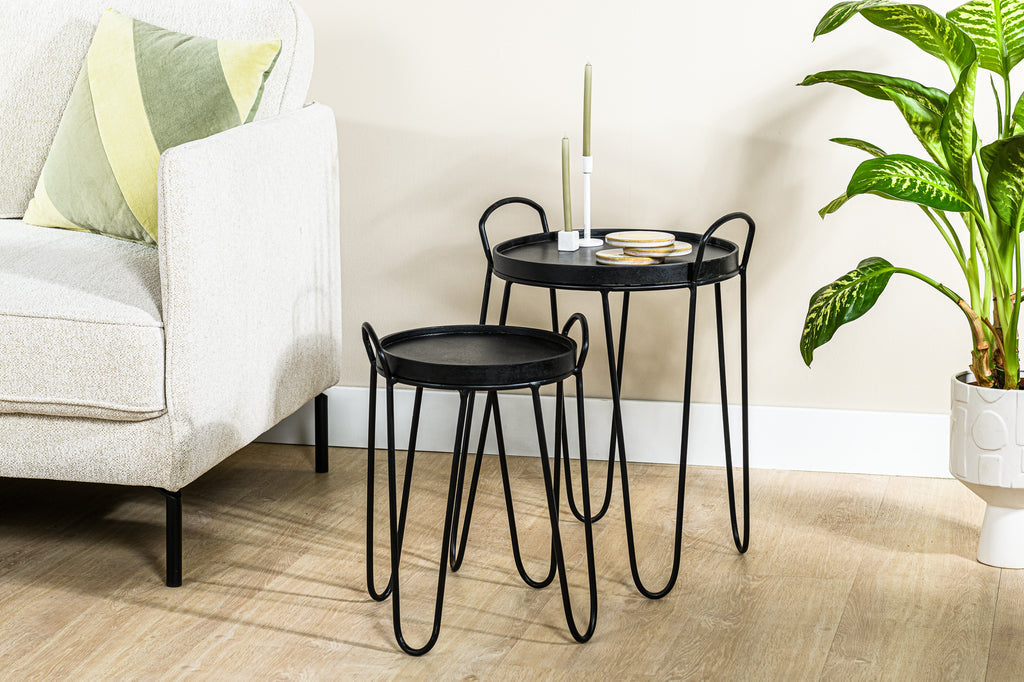 Beige bank met groen kussen, zwarte bijzettafels, groene kaars en potplant op houten vloer.