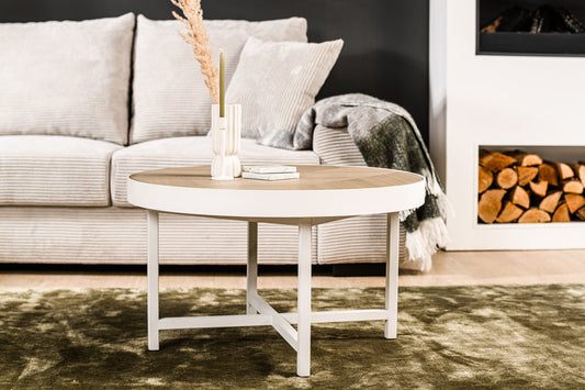 Ronde salontafel met vaas, droogbloemen en boek, voor een beige bank met kussens en grijze plaid.