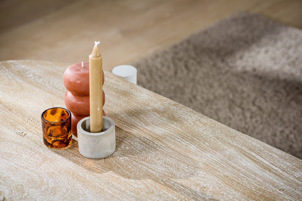 Beige kaars in stenen houder, rode kaarsen en amber glazen houder op licht houten tafel, bruine mat.