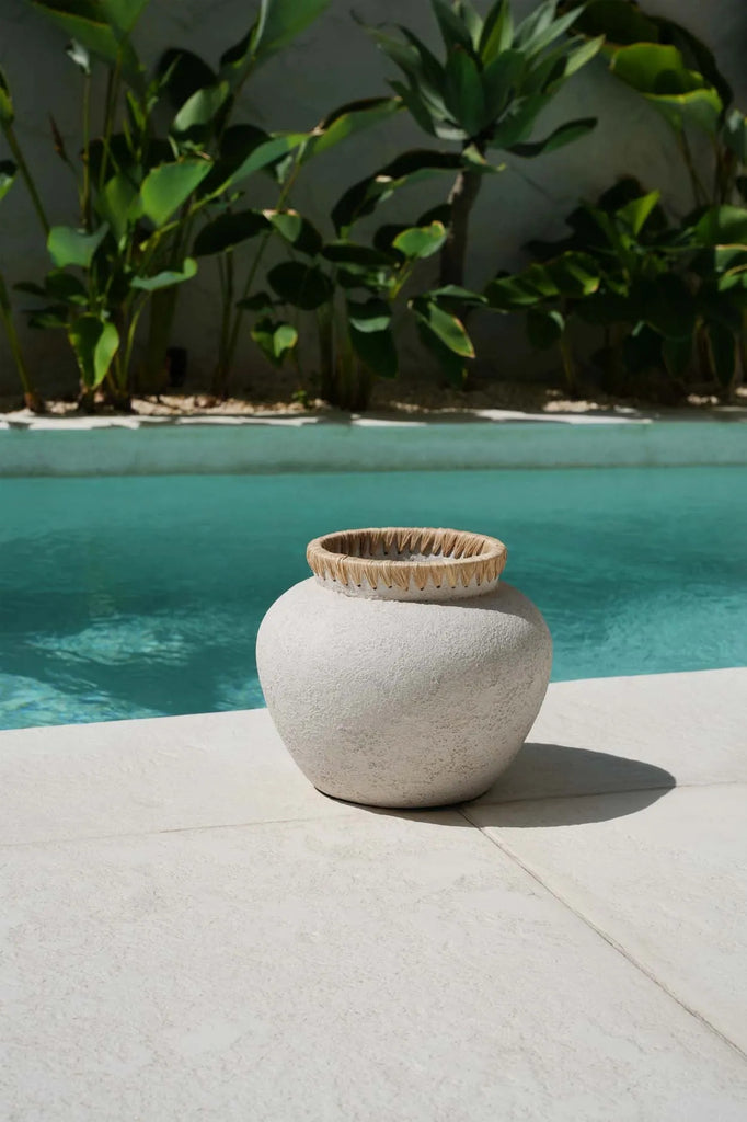 Zandkleurige keramische vaas op terras, omgeven door tropische planten en turquoise zwembad onder zonlicht.