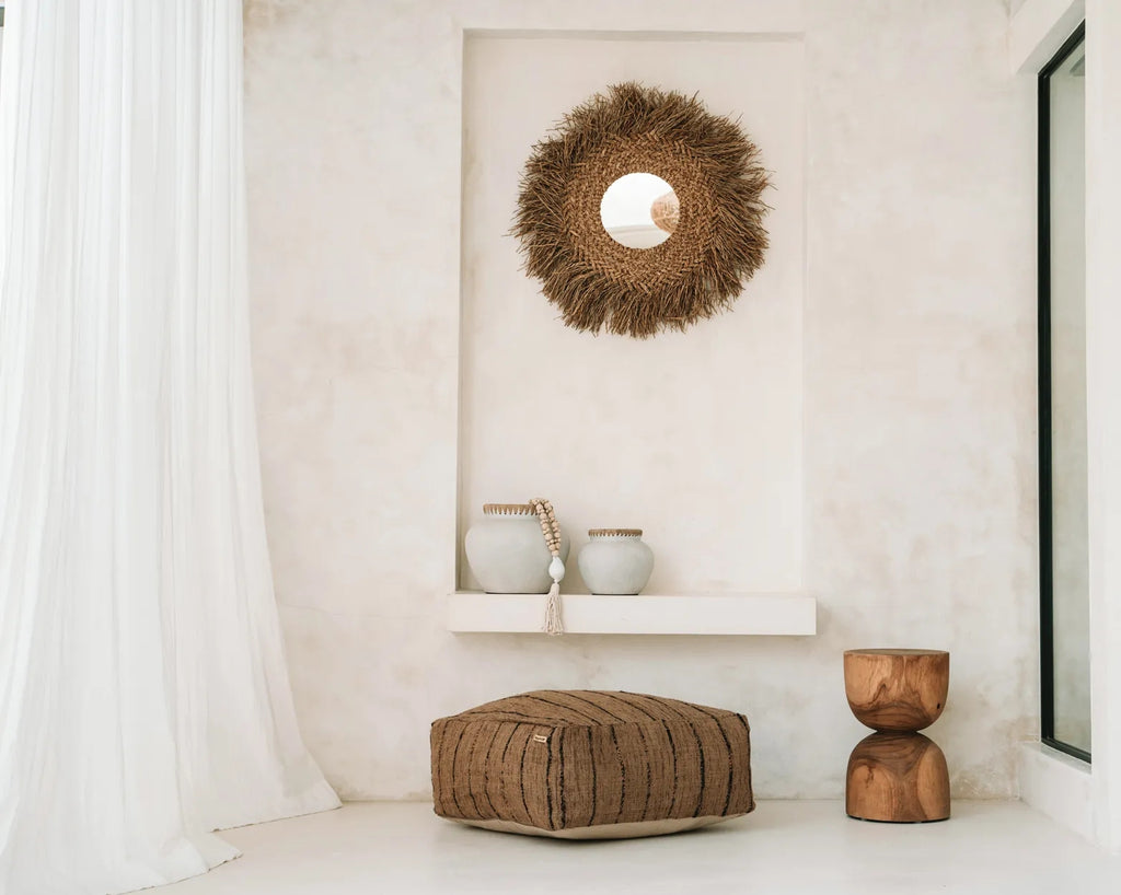 Stijlvolle boho-hoek met rieten spiegel, beige poef, houten kruk en vazen op een lichte achtergrond.
