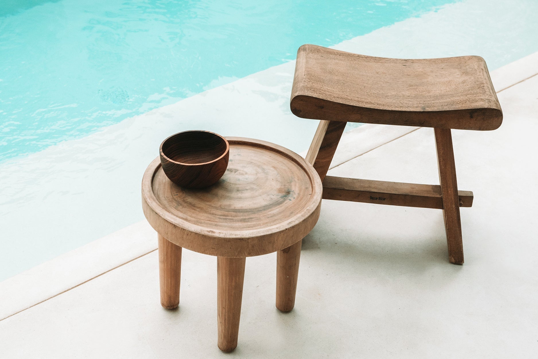 Houten kruk en tafel met een kom, naast een helderblauw zwembad, op een minimalistisch wit terras.