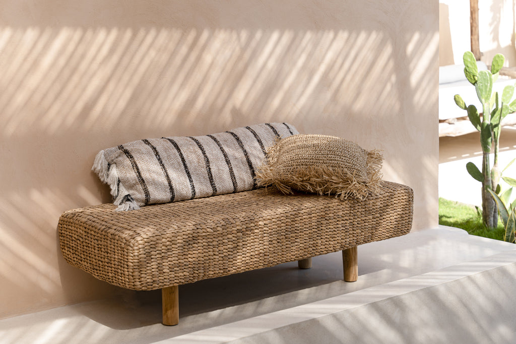 Rotan bank met gestreept kussen en geweven kussen in zonlicht, zachte schaduwen, beige muur, cactus op achtergrond.