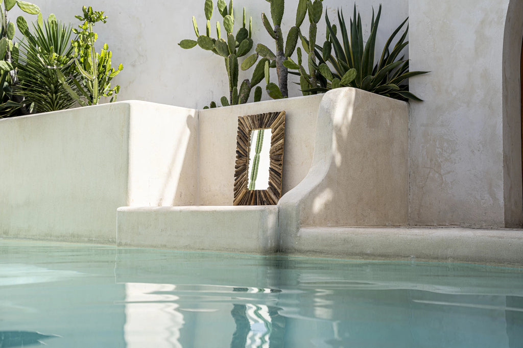 Spiegel met houten lijst aan zwembad bij minimalistische muur, omringd door tropische planten en cactussen.