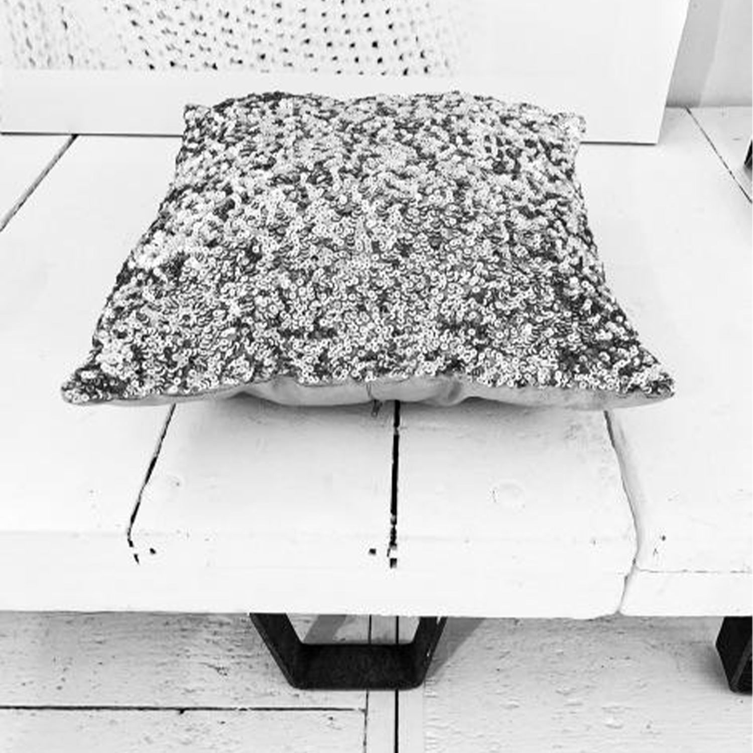 Glitterend sierkussen met pailletten ligt op een rustieke, wit geschilderde houten plank in een monochrome setting.