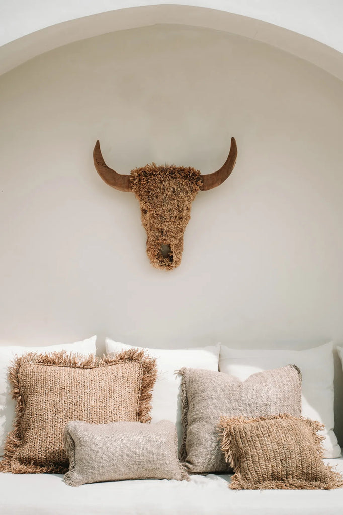 Buffelkopdecoratie aan muur boven een bank met beige kussens in verschillende maten, rustieke uitstraling.