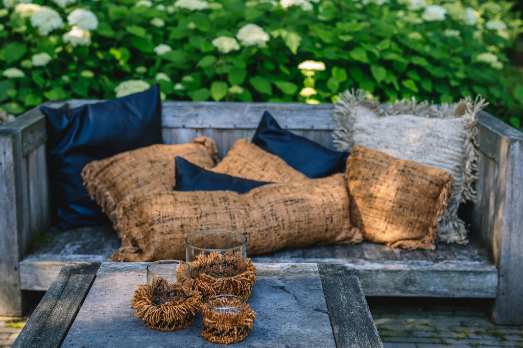 Houten bank in tuin met bruine en blauwe kussens, omgeven door groene planten. Rustieke glazen op tafel.