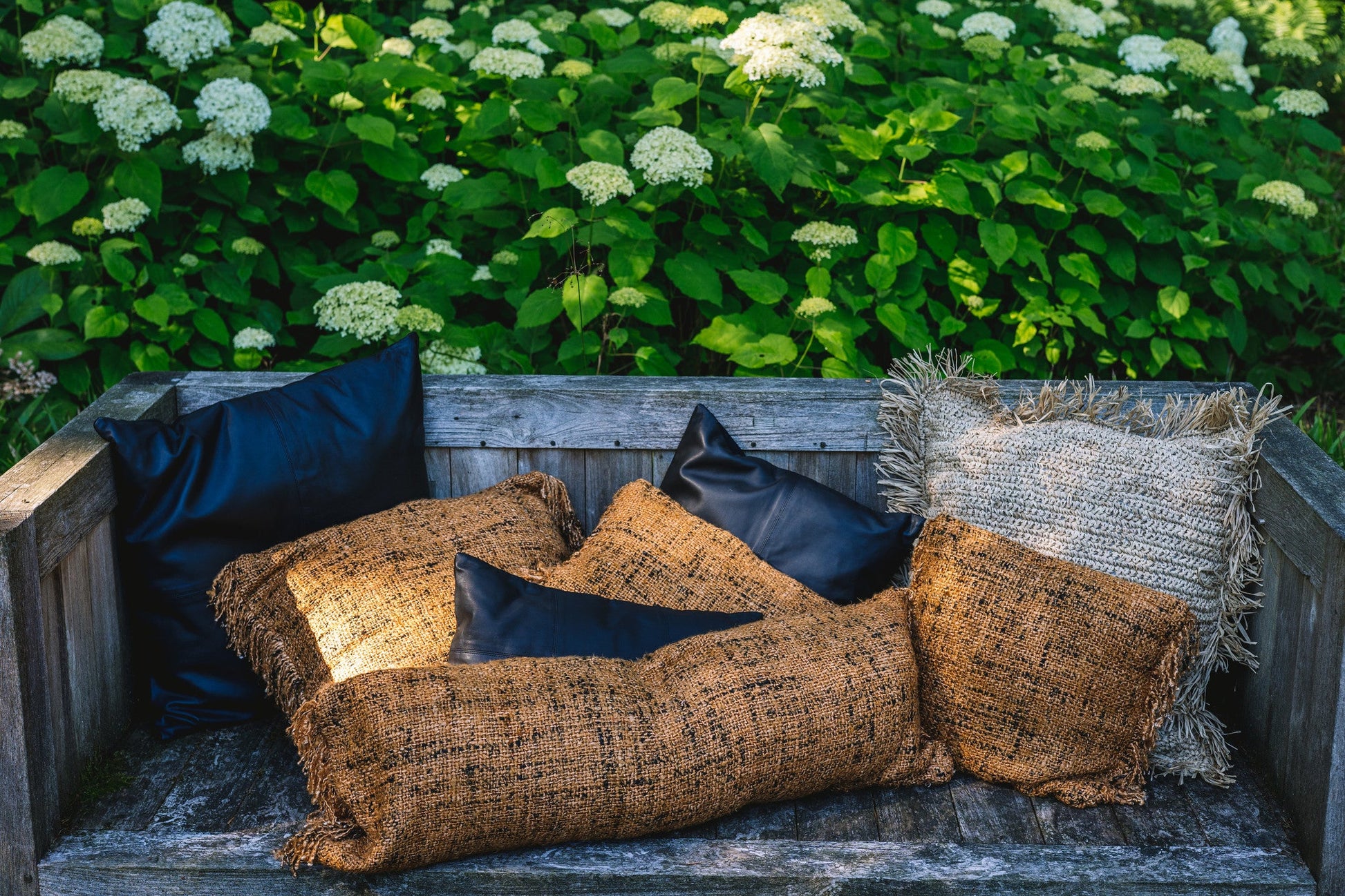 Houten bank met rieten en zwarte kussens in een tuin met groene bladeren en witte bloemen.