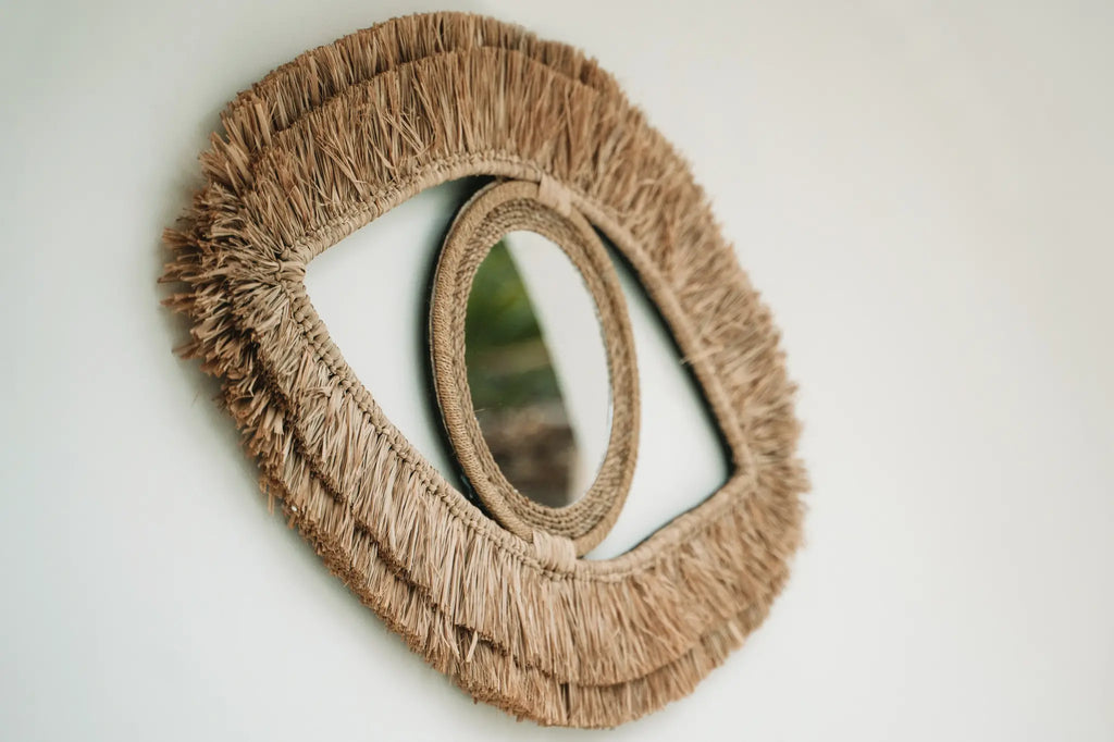 Ovaalvormige spiegel met rieten omlijsting die een oog imiteert, tegen een witte muur gemonteerd.