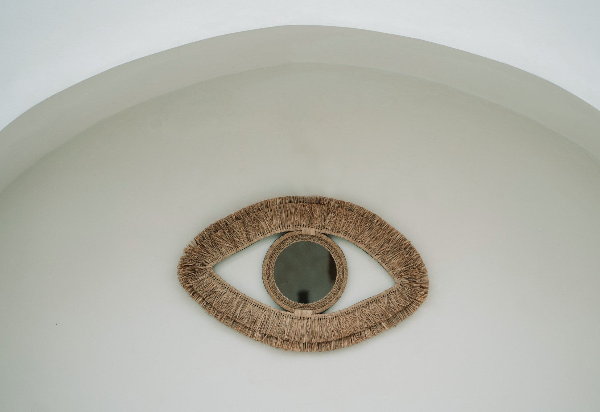Oogvormige rotan spiegel aan een witte wand, minimalistische en artistieke interieurdecoratie.