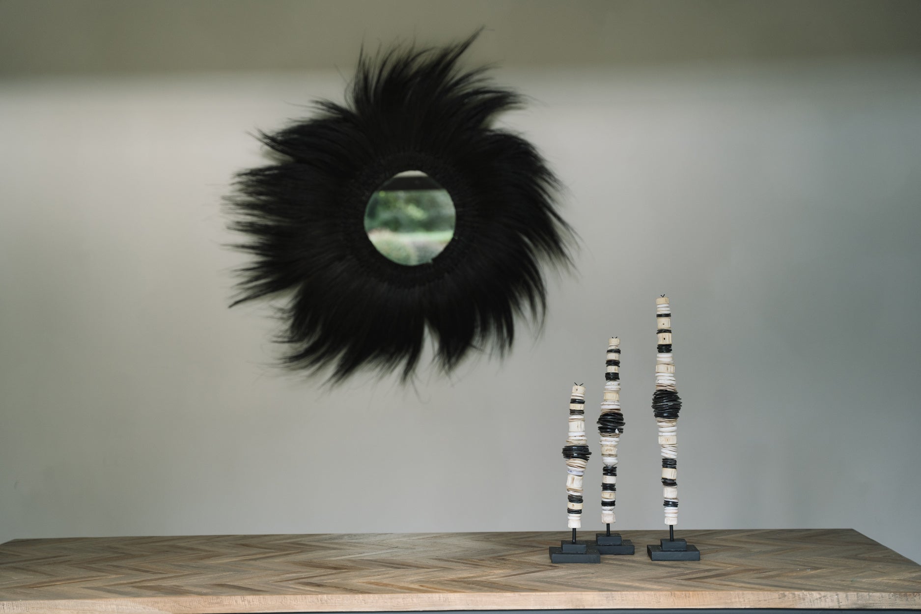 Zwart, rond kunstwerk met veren aan muur boven houten tafel met drie smalle, zwart-witte abstracte beeldjes.