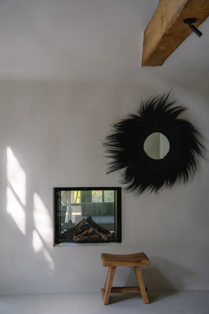 Houten kruk voor open haard met houtstapels, zwart decoratief ronde spiegel en rustieke houten plafondbalk.