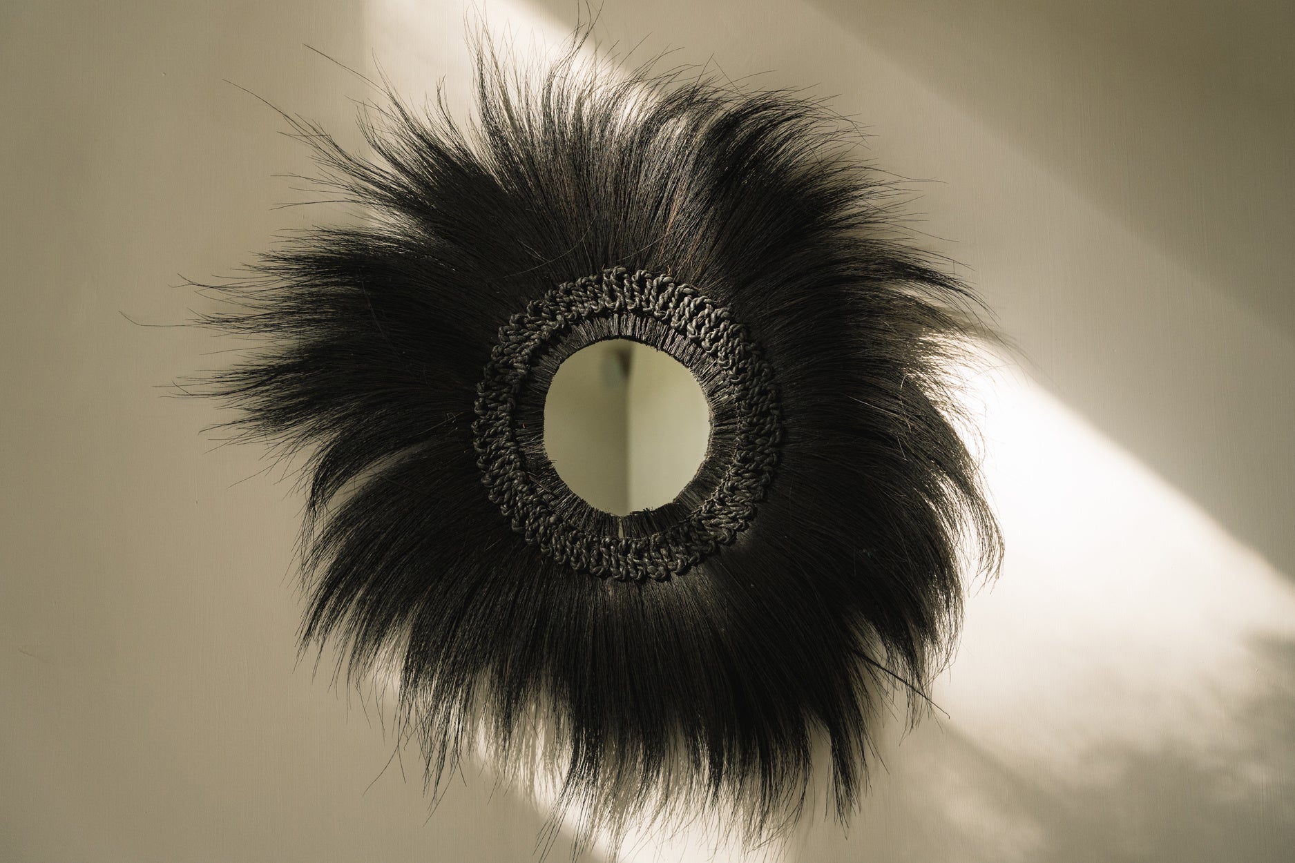 Rond decoratief object met spiegel, omgeven door zwarte vezels in een zonnestraal op een lichte muur.