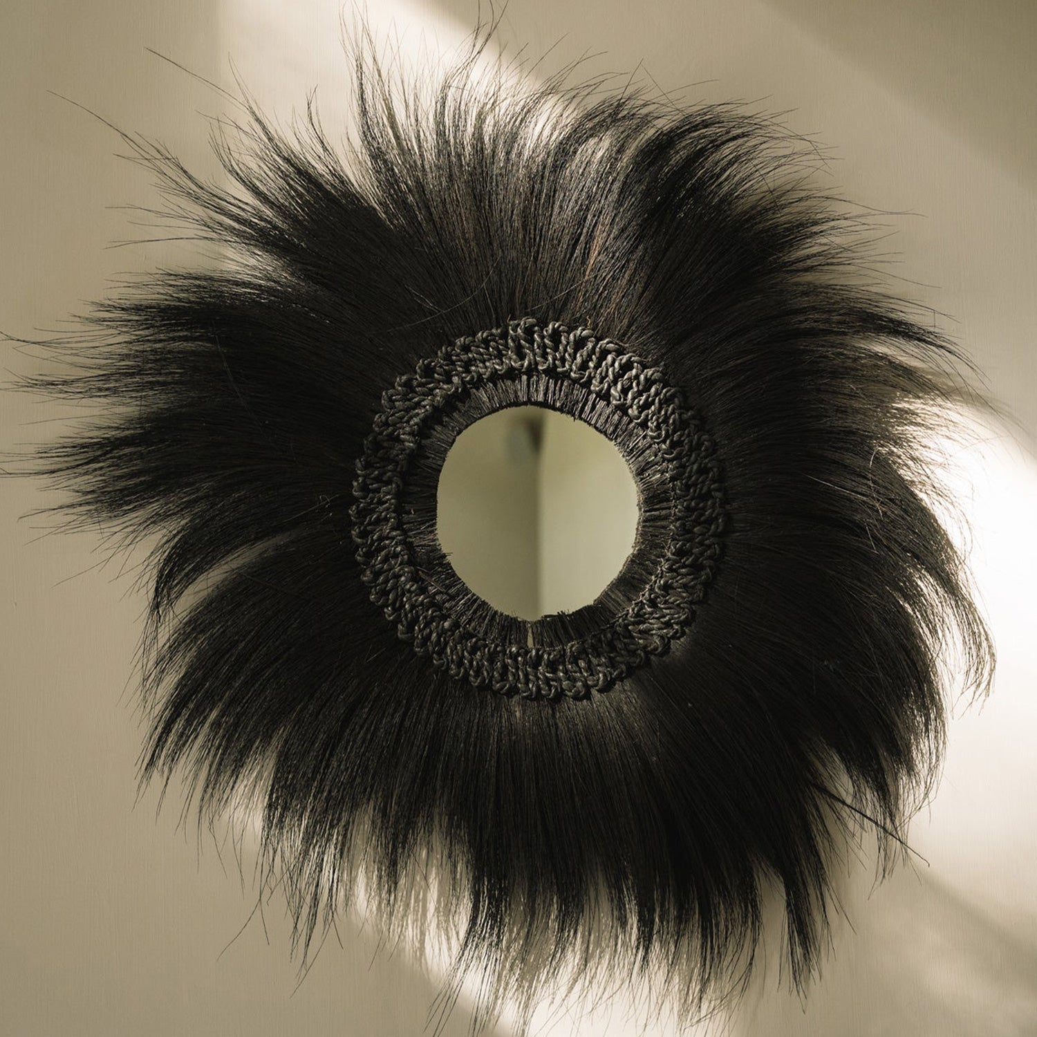 Rond decoratief spiegelontwerp met donker vlechtwerk en uitgespreide zwarte vezels tegen een lichte muur.
