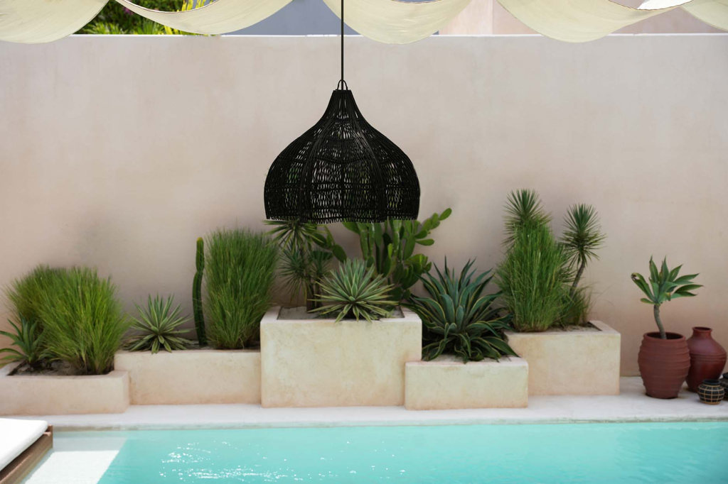 Zwart gevlochten lamp boven zwembad, omringd door groen struikgewas en terracotta potten tegen een beige muur.