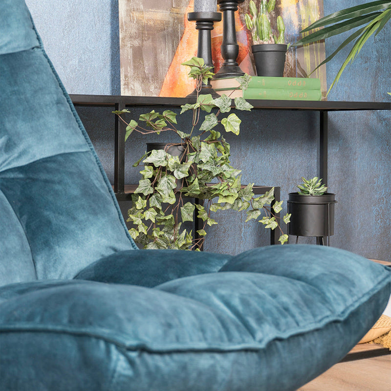 Blauw fluwelen stoel voor een tafel met groene planten, boeken en zwarte kandelaars tegen blauwe muur.