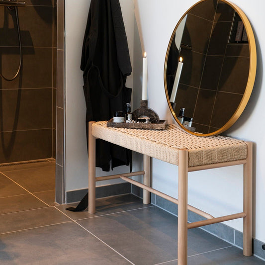 Ronde spiegel met gouden rand op houten bankje, brandende kaars en verzorgingsproducten in moderne badkamer.