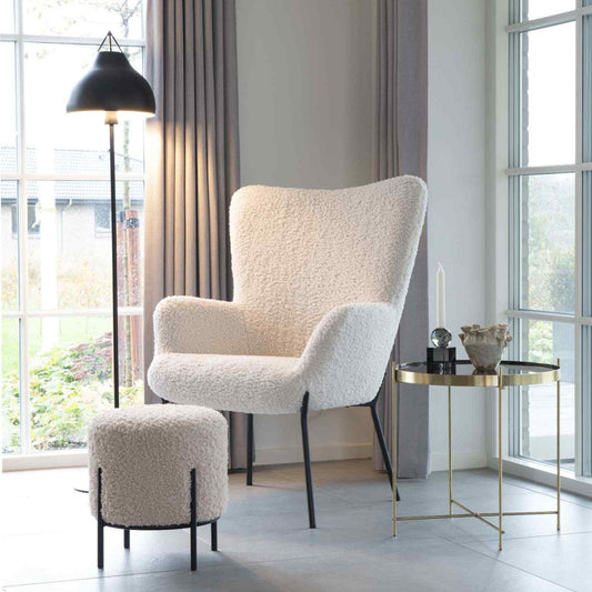 Warme woonkamer met een crèmekleurige fauteuil, bijpassende poef, gouden bijzettafel en vloerlamp.