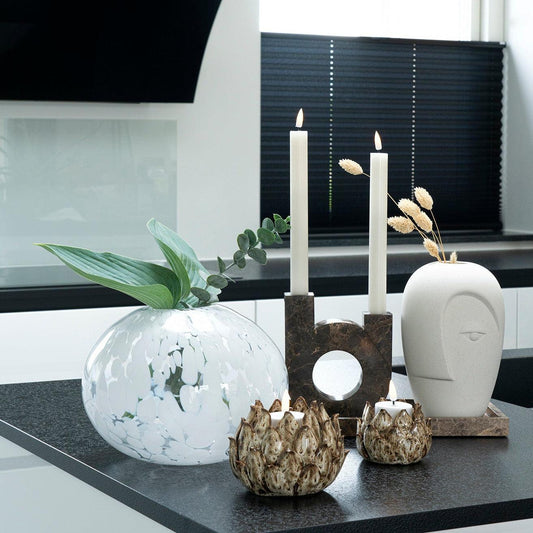 Stijlvolle compositie van een glazen vaas met bladeren, sculpturaal kaarsenhoudertje en moderne gezichtsvorm.