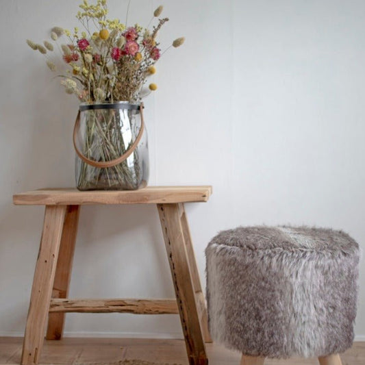 Glazen vaas met droogbloemen op houten kruk, naast een grijs pluchen voetenbankje in een minimalistisch interieur.