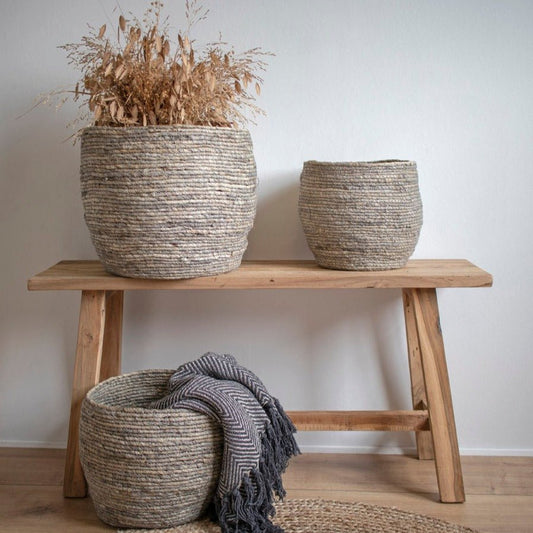 Grof geweven manden op houten bank, gevuld met gedroogde grassen en grijs geribbelde plaid, rustiek ingericht.