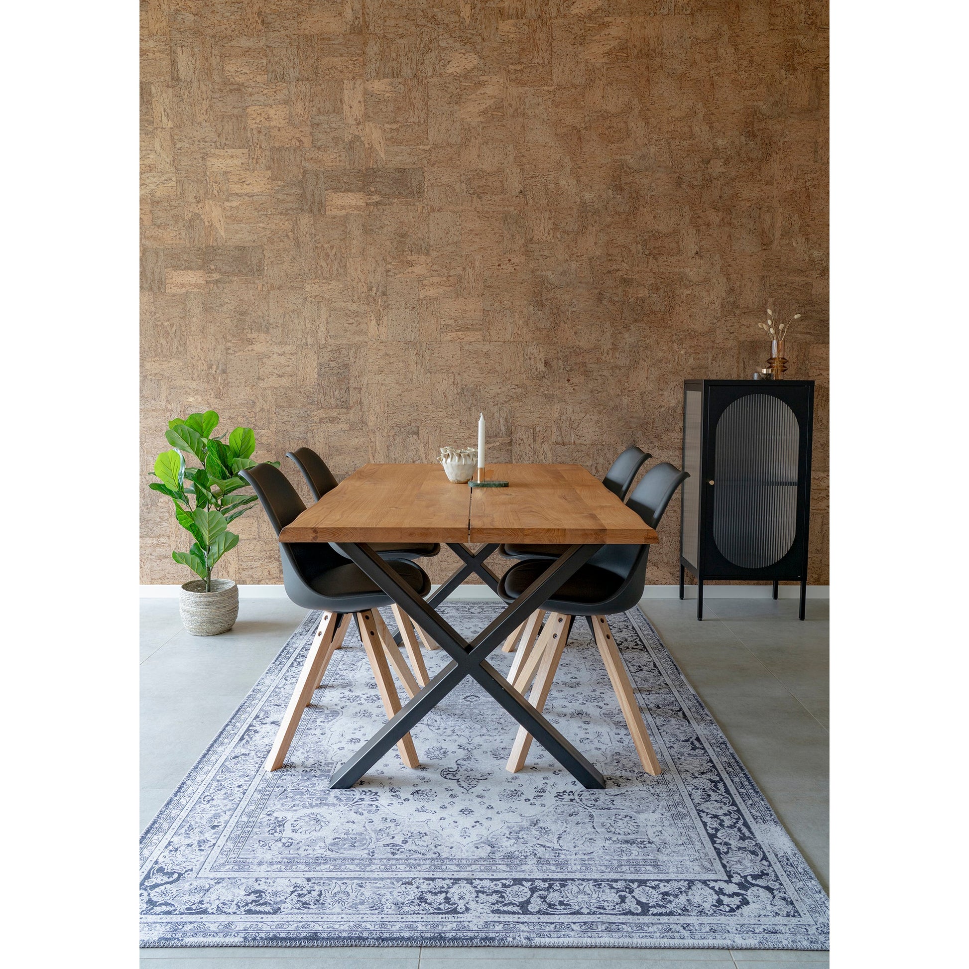 Eettafel met houten blad en zwarte stoelen op sierlijk tapijt, naast groene plant en zwart kastje.