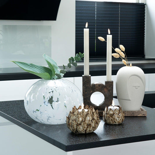 Witte keramische vaas met blad, modern kunstbeeld, kaarsen, en decoratieve kandelaars op zwart aanrecht.