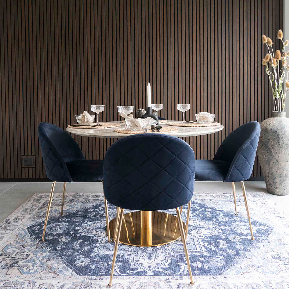 Elegante eethoek met ronde tafel, blauwe stoelen, kristallen glazen, en vintage tapijt op houten vloer.
