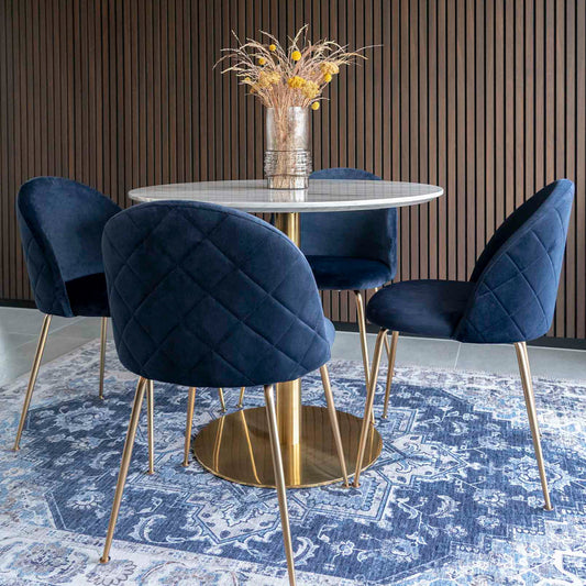 Ronde eettafel met witte marmeren blad, blauw fluwelen stoelen op gouden poten en vaas met droogbloemen.