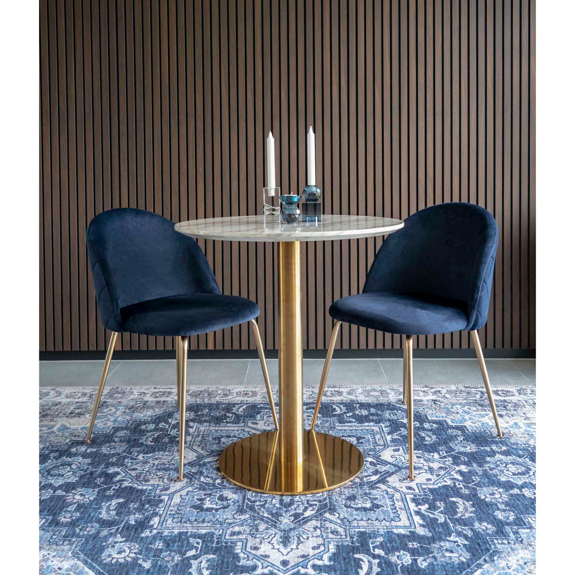 Ronde marmeren tafel met gouden poot, blauw fluwelen stoelen en kandelaars op Perzisch tapijt tegen houten wand.