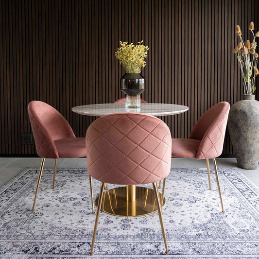 Roze stoelen rond een witte tafel met een vaas gele bloemen, op een sierlijk vloerkleed tegen een houten wand.