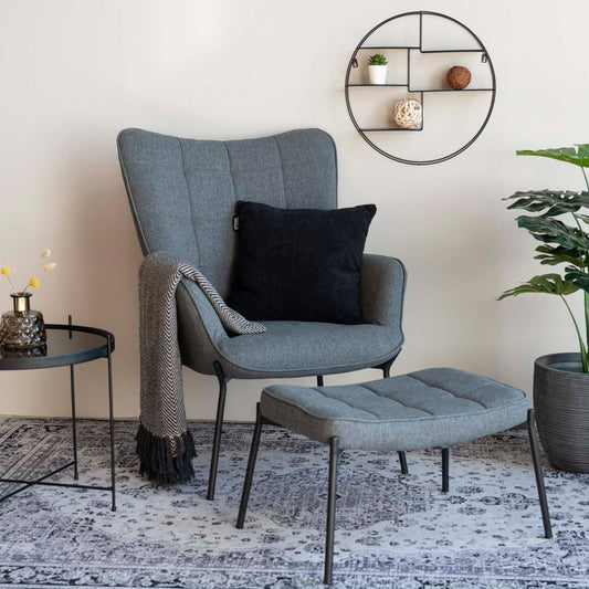 Grijze fauteuil met zwart kussen en plaid, bijzettafel met kaars, rond wandrek en groene plant op oosters tapijt.