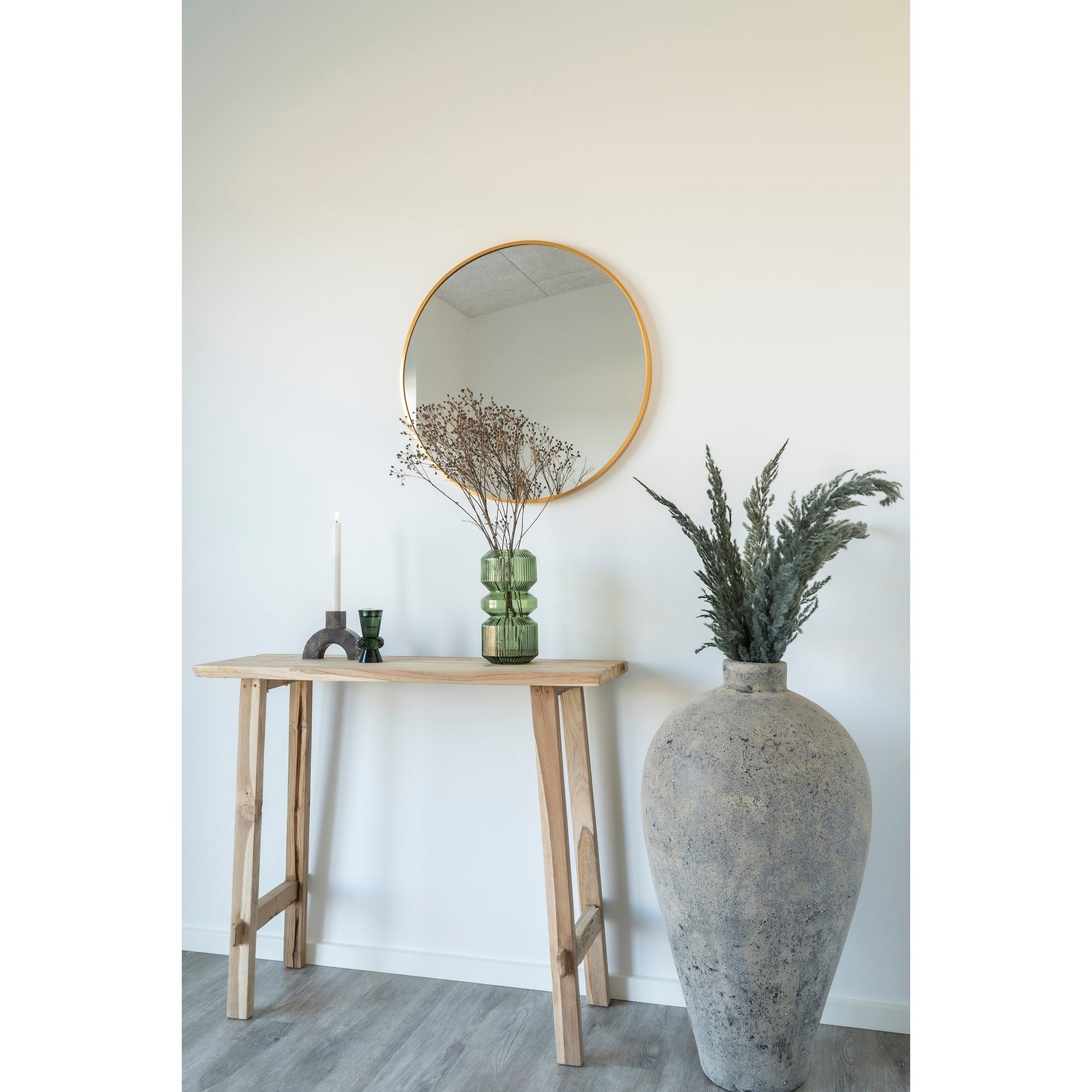 Rond gouden spiegel boven houten tafel met groene vaas, kaars, en grote grijze pot met gedroogde takken.