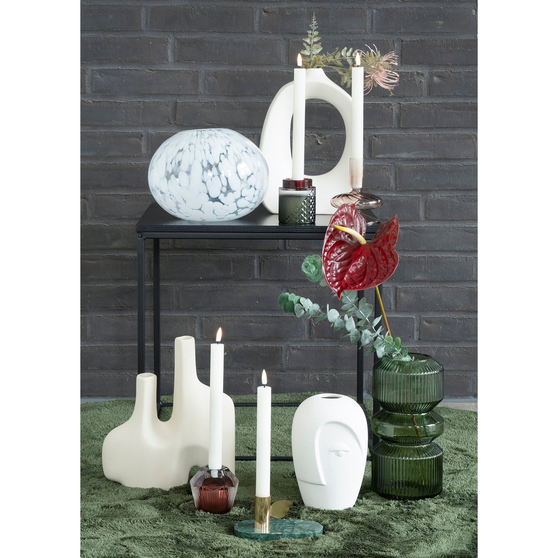 Stijlvolle display met marmeren vaas, witte kaarsen, modern keramiek en rode bloemen op groen vloerkleed.