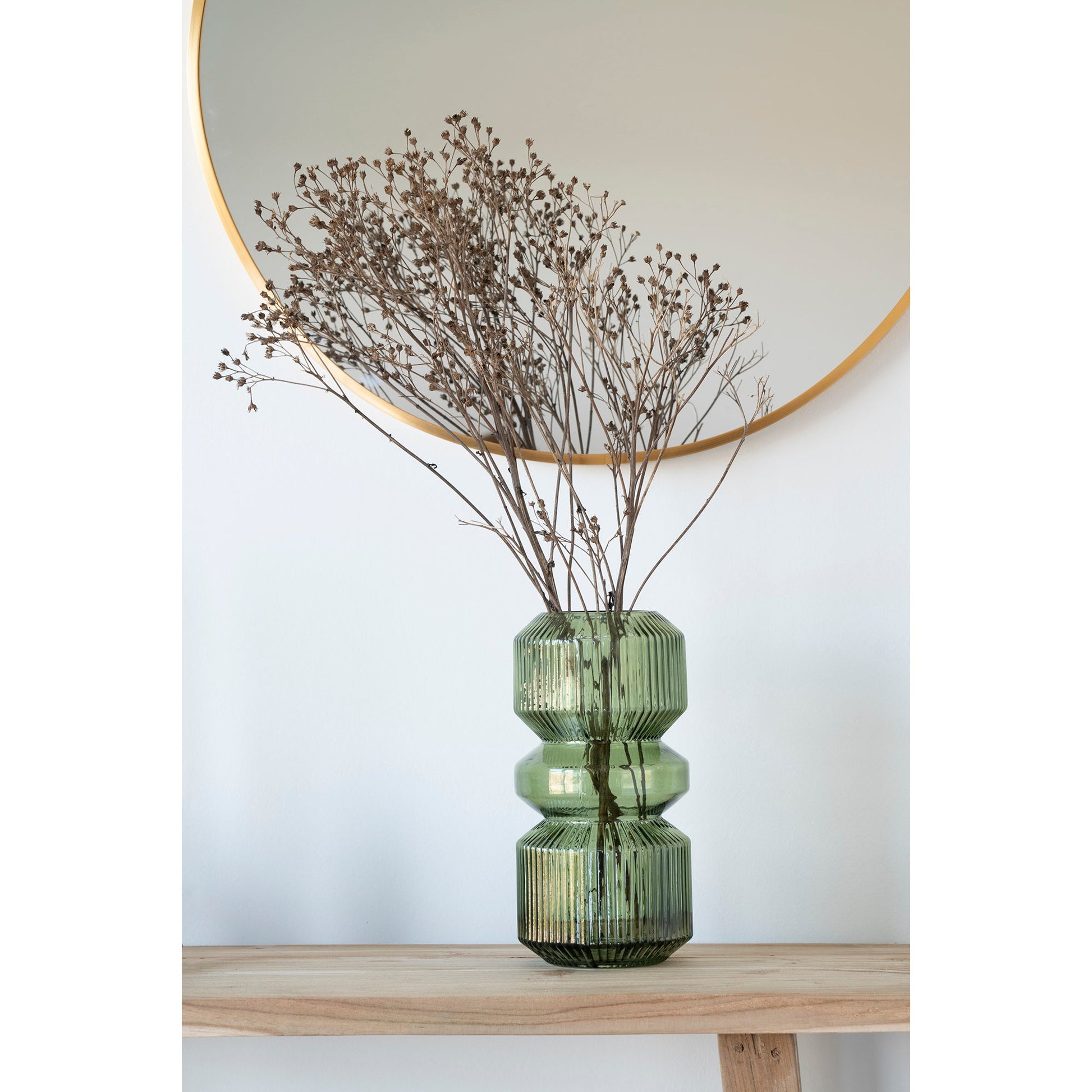 Groene glazen vaas met gedroogde takken op houten plank, tegen witte achtergrond en ronde spiegel.