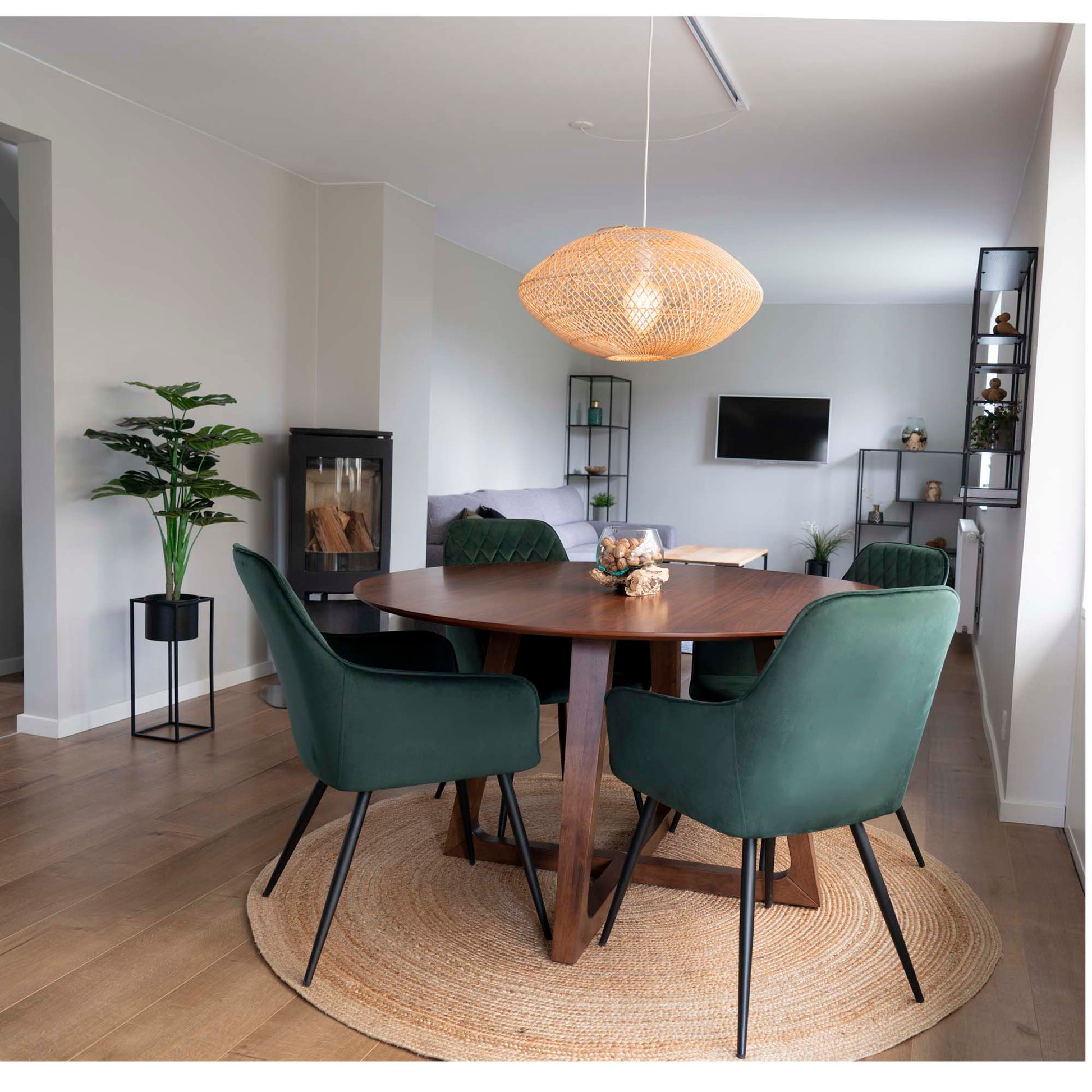 Moderne eetkamer met rond houten tafel, groene stoelen, rieten lamp en plant in lichte, minimalistische ruimte.