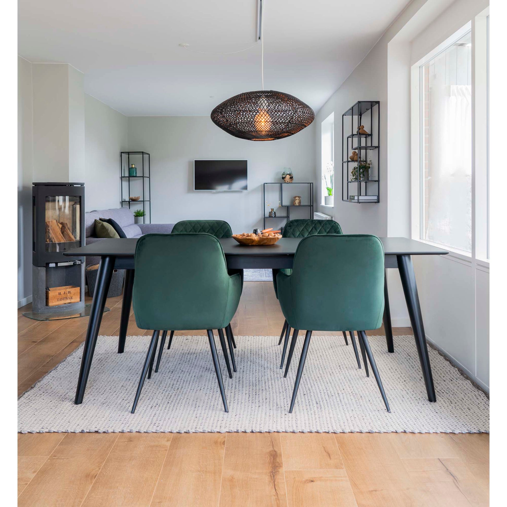 Modern eetkamer met zwarte tafel, groene stoelen, houten vloer en hanglamp, met uitzicht op woonkamer.