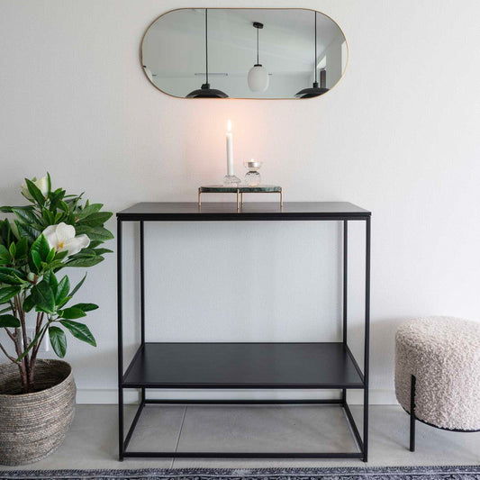 Strakke zwarte console met brandende kaars, ronde spiegel erboven, plant links en witte poef rechts.