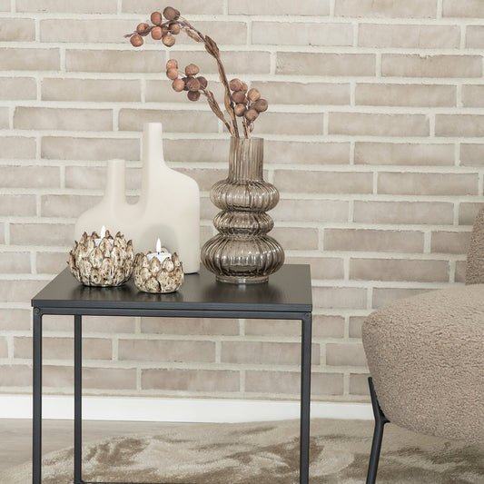 Glazen vaas met gedroogde takken, keramische vazen en kaarsen op zwart bijzettafeltje voor lichte bakstenen muur.