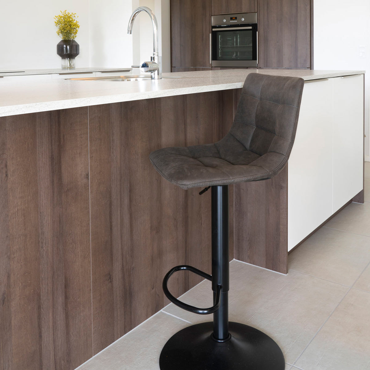 Donkerbruine barkruk met metalen voet bij modern keukenblad van hout en marmer, vaas met gele bloemen.