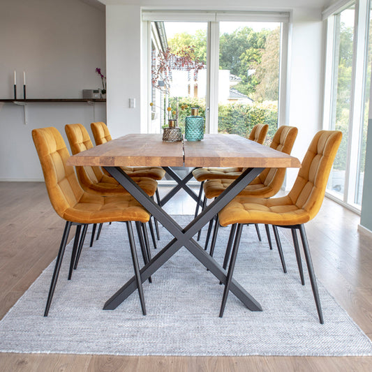 Houten tafel met X-vormige poten, omringd door acht okergele stoelen in een lichte eetkamer met grote ramen.
