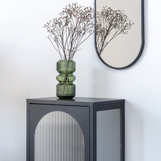 Donkergroen glazen vaas met takken op zwart kastje naast een ovalen spiegel tegen een witte muur.