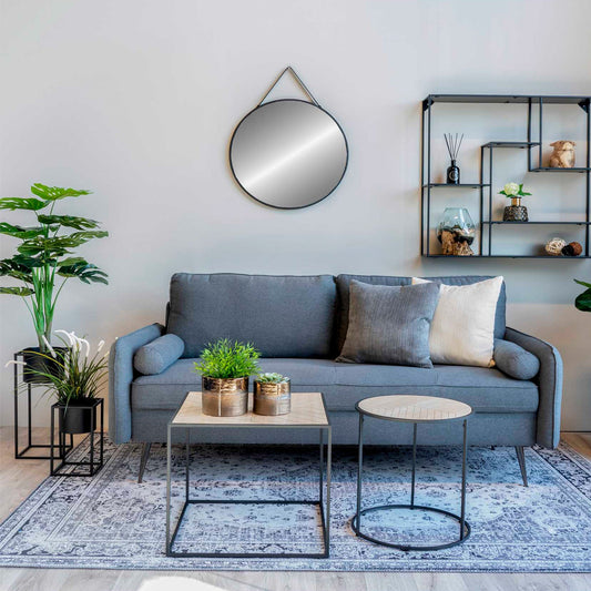 Stijlvolle woonkamer met grijze bank, ronde spiegel, metalen planken, groen planten en moderne tafels.