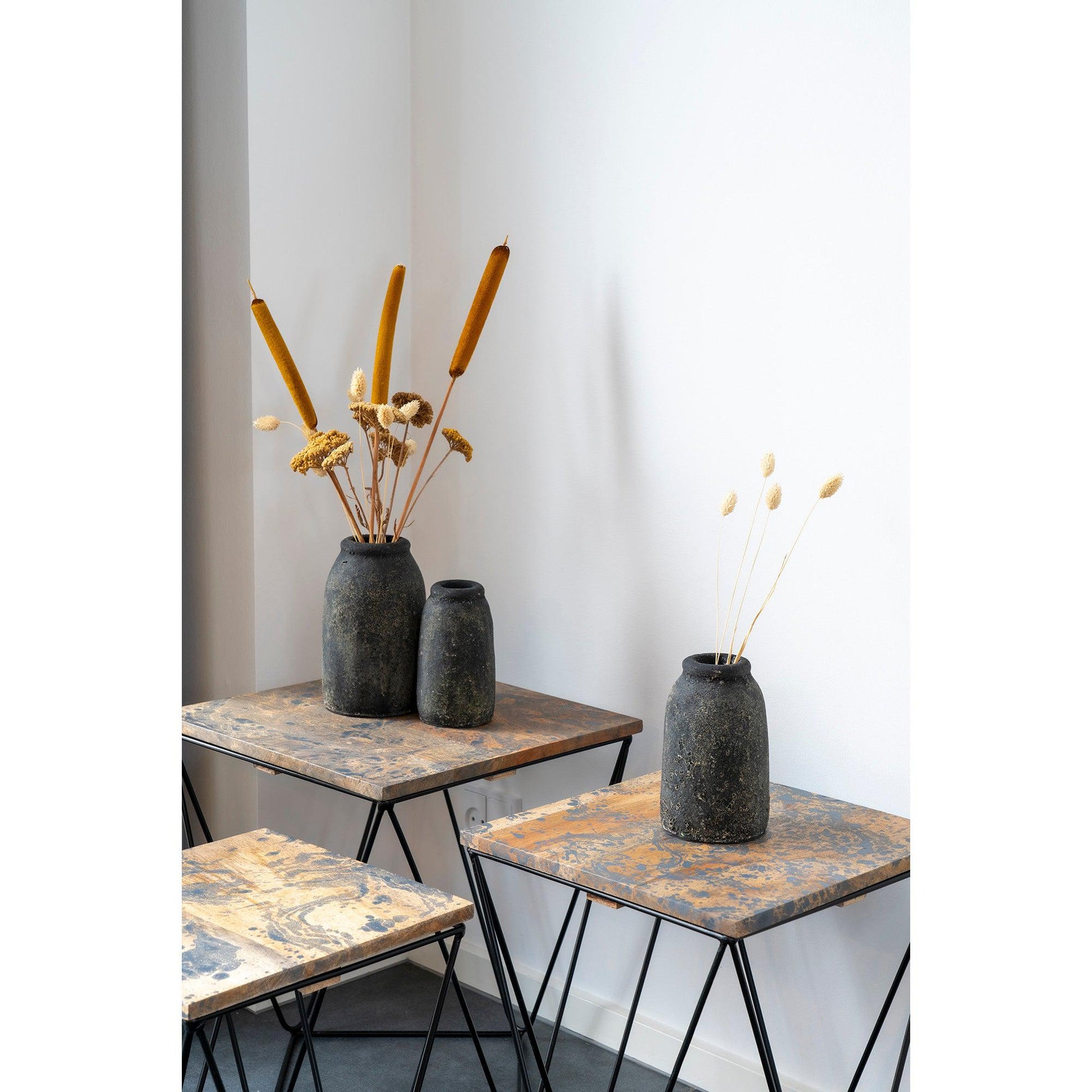 Donkere, rustieke vazen met rietpluimen en droge bloemen op geometrische houten tafels tegen witte muur.