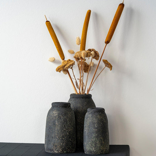 Drie ruwe, zwarte vazen met gedroogde rietpluimen en bloemen tegen een witte muur.