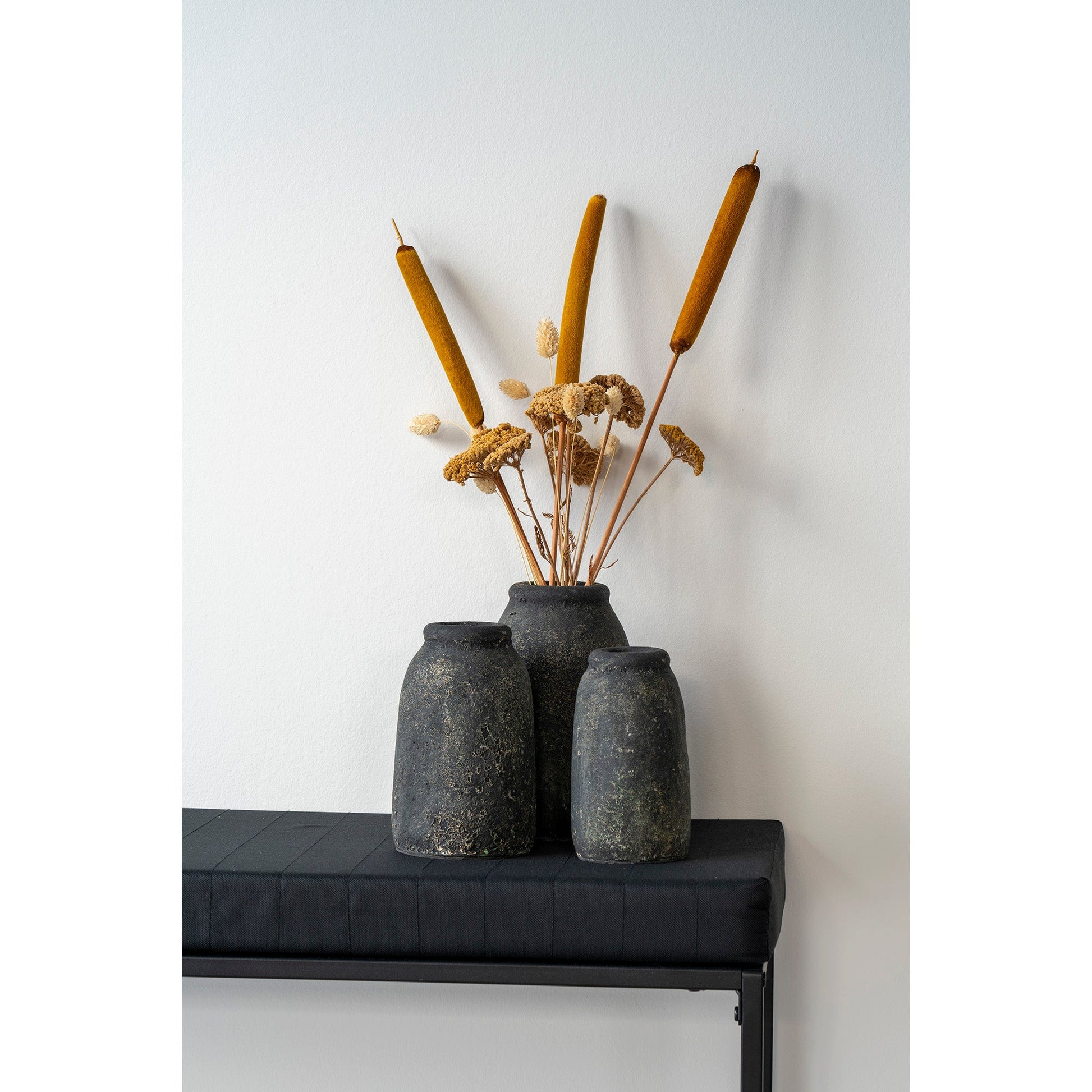 Drie rustieke donkere vazen met gedroogde bloemen en biezen op een eenvoudige zwarte console, minimalistisch design.