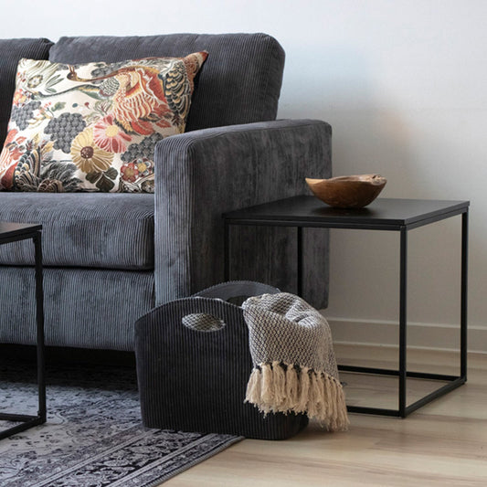 Grijze ribfluwelen bank met bloemenkussen, bijzettafel met houten kom en mand met geruite deken.