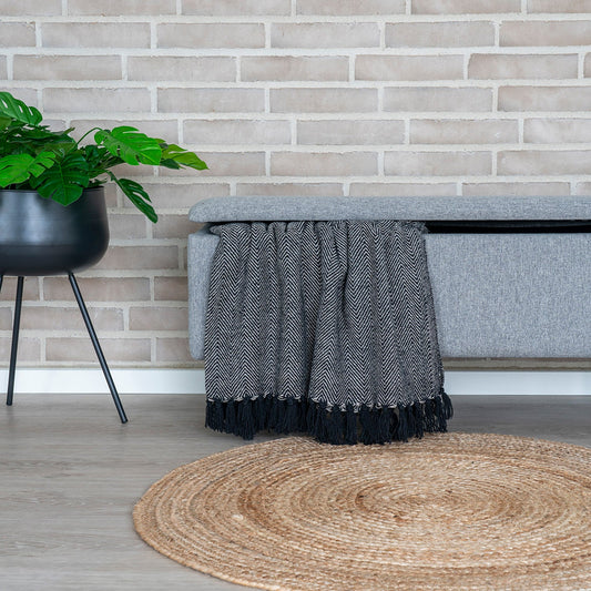 Grijze bank met zwart-wit visgraatkleed, zwarte plant op pot, jute vloerkleed en bakstenen muur.