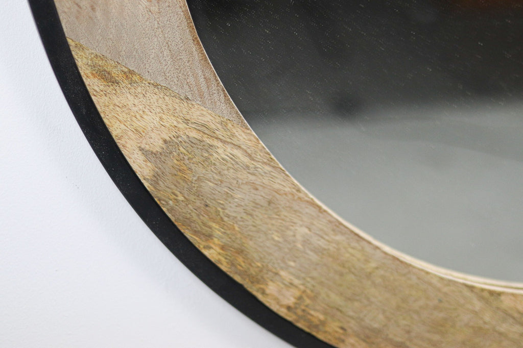 Detailopname van een ronde spiegel met een houten rand en zwarte afwerking tegen een witte muur.