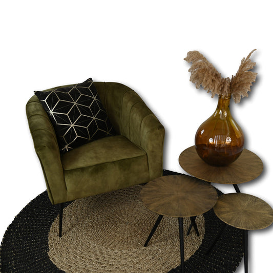 Groene fluwelen fauteuil met geometrisch kussen naast houten tafels en amberkleurige vaas op rond tapijt.